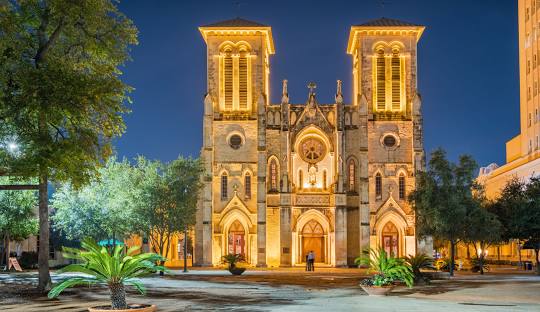 A scene of the front of San Fernando Cathedral - Night Time with beautiful lighted accents with a couple standing in front.  Palm trees line the courtyard.